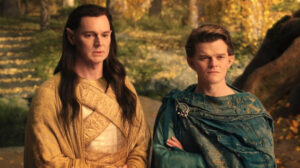 Benjamin Walker as Gil-galad and Robert Aramayo as Elrond in The Rings Of Power, standing side-by-side in an autumnal forest glade. Gil-galad has long brown hair worn loose, and wears a shimmering gold mantle and gold fabric bandolier. Elrond has short brown hair, and wears a dusty-green cloak and long-sleeved tunic.