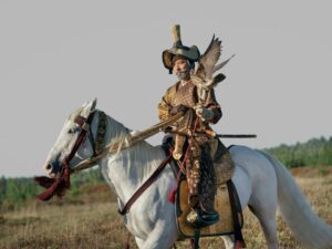 Hiroyuki Sanada as Yoshii Toranaga in Shogun, seated on a white horse on a grassy hillside, with a hawk perched on his hand. He is wearing golden-brown garments, and a tall straw hat with a chinstrap.