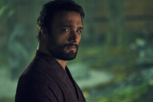 Cosmo Jarvis as John Blackthorne in Shogun, from the chest up, wearing a dark brown robe. He has close-cropped brown hair and a beard.