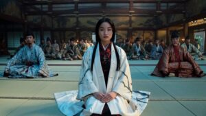 Anna Sawai as Toda Mariko in Shogun, kneeling on straw-mats covering the floor of a large room. She is wearing a white robe over a black and blood-red kimono and has a crucifix on a necklace. She has long black hair, parted in the middle. There are people seated behind her and to either side.