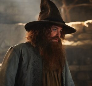 Rory Kinnear as Tom Bombadil in The Rings Of Power, close-up. He has long curly reddish-brown hair and a beard. He is wearing a tall brown hat with a dark blue feather through it, and a blue-gray coat over a tunic of indeterminate color with a brown leather belt.