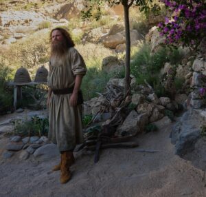 Rory Kinnear as Tom Bombadil, standing outside in a sandy garden of cacti, with beehives, lemon-trees, and pink flowers climbing the stone walls of his house. He has long reddish-brown hair and a beard, and is wearing a white tunic with rolled-up sleeves and yellow boots.