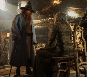 Rory Kinnear as Tom Bombadil, standing in front of Daniel Weyman as the Stranger, who is sitting in a wooden chair. Tom has long reddish-brown hair and a beard. He is wearing a tall brown hat and a blue coat over a tunic with a brown leather belt and yellow boots, carrying a wooden walking-stick. The Stranger has long gray hair and is wearing a moss-colored robe. They are in Tom's house, which has star-charts etched on the stone ceilings and rugged furniture.