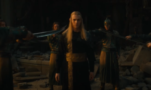 Charlie Vickers as Sauron in The Rings Of Power, wearing a black long-sleeved robe with ornate gold embroidery and a wide gold belt, standing surrounded by long-haired Elven warriors wearing armor and holding swords to his throat.  He has long blond hair.