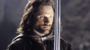 Viggo Mortensen as Aragorn, brandishing the sword Anduril in front of his face. He has shoulder-length dark brown hair, and wears a gray cloak over brown garments.