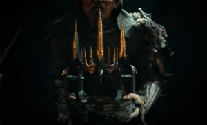 An orc dressed in black armor carrying a slab of polished black stone, upon which sits a black iron tiara with dagger-like prongs that catch the firelight from nearby torches and glow reddish-orange.