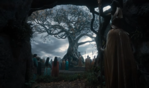 A bare and leafless tree under a cloudy sky, surrounded by Elves in colorful robes, and guarded by soldiers wearing winged helmets and gold cloaks, carrying spears.