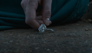 Nenya, a ring of braided silver with a large white gemstone set in it, lies on the ground at the foot of a person wearing a floor-length blue-green gown, who is stooping to pick up the ring in their left hand.