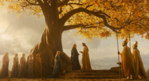 Wide shot of a round stone dais in the shade of a tall tree with golden leaves. Ben Daniels as Cirdan and Benjamin Walker as Gil-galad stand upon the dais, while Morfydd Clark as Galadriel ascends the dais by a flight of short steps from the left. Cirdan has long gray hair and wears a gray robe. Gil-galad has long dark hair, a crown of golden laurel leaves, and wears a golden robe. Galadriel has long blonde hair and wears a green gown. Veiled attendants and soldiers wearing gold cloaks and winged helmets, wielding spears, stand nearby.