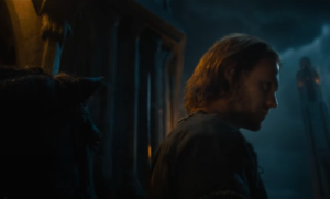 In the foreground, Charlie Vickers as Halbrand, in profile, stands next to a horse. It is nighttime, and raining. Behind him are tall towers. He has shoulder-length brown hair and wears a brown cloak.