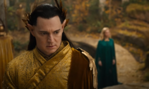 Benjamin Walker as Gil-galad, standing in the foreground, while Morfydd Clark as Galadriel stands behind him, out-of-focus. Gil-galad has long dark brown hair, and wears a circlet of golden laurel leaves and a golden-brown cape over the left shoulder of his gold robe. Galadriel has long blonde hair, and wears a floor-length blue-green gown.