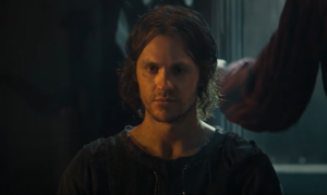 Charlie Vickers as Halbrand, sopping-wet, standing in a doorway, while behind him, Charles Edwards as Celebrimbor closes a door to shut out the rain. Halbrand has shoulder-length brown hair and wears a brown tunic.