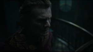 Charles Edwards as Celebrimbor, in close-up, standing on a mezzanine overlooking his forge. It is dark. His face is lined with worry and possibly fear. He has short brown hair, and wears a red robe with gold embroidery.