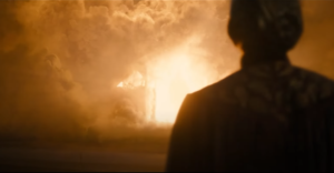 Charles Edwards as Celebrimbor, from behind, approaching a white light burning through a barrier of fog and smoke. He has short brown hair and wears a dark red robe with gold embroidery.