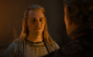 Charlie Vickers as Annatar in The Rings Of Power, facing Charles Edwards as Celebrimbor, whose back is turned to the camera. Annatar has long blonde hair held back by a golden circlet, and wears a white mantle over a white tunic with silver embroidery. Celebrimbor has short brown hair and wears a dark red robe with gold embroidery.