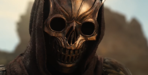 Close-up image of a person of indeterminate age and gender, wearing a brown hood over an ornate bronze mask shaped into a leering skull with gaping eyesockets.