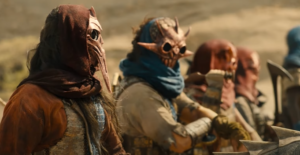 A row of people of indeterminate age and gender on horseback, wearing red and blue scarves over gold and bronze masks shaped into demonic faces with beaks and horns.