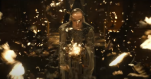 A woman with slicked-back white-blond hair, wearing a white hood and silver armor over a white gown, standing in a temple. In her hands is a growing ball of light, and a swarm of brown butterflies explodes outward from the light, fluttering all around her.