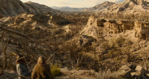 In the foreground, Rory Kinnear as Tom Bombadil and Daniel Weyman as The Stranger stand upon a cliff, looking out over a dead forest of brittle-branched, leafless trees in a rugged mountain valley. Tom wears a tall blue hat and a blue coat. The Stranger has long gray hair and wears a mossy green robe.