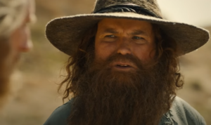 Rory Kinnear as Tom Bombadil, in close-up. He has long curly reddish-brown hair and a bushy beard. He wears a tall blue hat and a blue coat.