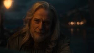 Ben Daniels as Cirdan in close-up. He has long gray hair, slightly curly, and a gray beard. He wears a gray robe with golden waves embroidered around the collar.