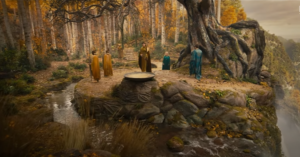 A cliffside in Lindon, upon the edge of which grows a tall mallorn tree with golden leaves. Within its shade sits a dais, encircled by a stream falling off the edge of the cliff, and upon that dais stands a basin filled with water. Benjamin Walker as Gil-galad is approaching the basin. He has long dark hair and wears a golden robe. Nearby stands Robert Aramayo as Elrond. He has short brown hair and wears a blue robe. In the background stand two Elven soldiers in gold cloaks, wielding spears, and Morfydd Clark as Galadriel. She has long blond hair and wears a green mantle.