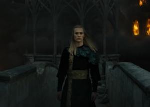 Charlie Vickers as Sauron, striding down a broken stone walkway in Eregion, while behind him, buildings collapse and flames erupt as they fall. He has long blonde hair, and wears a cape of glossy black feathers or scales over a black long-sleeved robe with gold embroidery down the front and a wide gold belt.