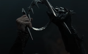 A pair of hands, belonging to Sam Hazeldine as Adar, holding up a black iron crown with dagger-like prongs. One hand is encased in a thorny black gauntlet.