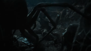 Maxim Baldry as Isildur, sprawled on the floor of a tunnel, pulling himself upright and wielding a knife. In the foreground, scuttling down from the ceiling, is a spider almost half his height, with long spindly legs. Isildur has shoulder-length shaggy brown hair and wears lightweight brown leather garments.