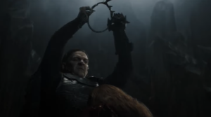 Sam Hazeldine as Adar, standing in front of a kneeling figure with long reddish-brown hair, holding aloft a black iron crown and preparing to bring it down heavily onto the person's head. He has long black hair, and wears black armor over a chain-mail tunic.