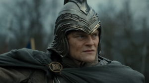 Robert Aramayo as Elrond, in close-up, wearing a silver helmet and a gray cloak with a gold brooch over silver armor.