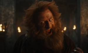 Owain Arthur as Durin IV, from the chest up, in the middle of a speech. He has red hair, and a bushy red beard. He wears rust-red armor over a red-and-gold tunic.