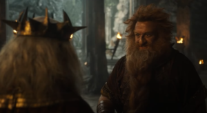 Owain Arthur as Durin IV, talking to Peter Mullan as Durin III, whose back is turned to the camera. Durin IV has red hair and a braided red beard. He wears rust-red armor over a red-and-gold short-sleeved tunic with leather gauntlets. Durin III has long white hair and wears an iron crown with short spikes. They are standing in an underground hall with stone pillars.