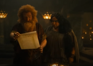 Owain Arthur as Durin IV, reading from a piece of parchment while Sophia Nomvete as Disa stands just behind him, reading over his shoulder. Durin has red hair and a bushy beard. He wears a red-and-gold short-sleeved tunic. Disa has long dark curly hair, and wears a loose gray gown with a cape.