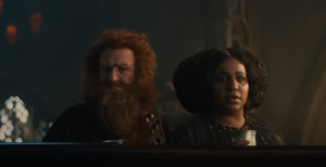 Owain Arthur as Durin IV and Sophia Nomvete as Disa, standing on the mezzanine overlooking Celebrimbor's forge. Durin has red hair and a bushy red beard. He wears red armor over a red-and-gold tunic. Disa has dark curly hair, piled up on the sides of her head. She wears a loose gray gown.