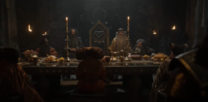 Peter Mullan as Durin III, presides over a banquet table, around which several Dwarven lords and ladies are seated. Durin has long gray hair and a longer beard, and wears a spiky iron crown and a gold robe.