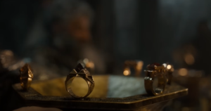 Close-up shot of a gold chalice on a table, around the rim of which are placed seven gold rings, each standing upright and crowned with a heavy jewel.