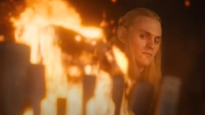 Charlie Vickers as Sauron, looking into the flames of a brazier standing in the foreground, which have taken the shape of a Balrog with curled horns, glowing eyes, and a gaping maw. Sauron has long blond hair, and wears the faintest of smirks on his face.