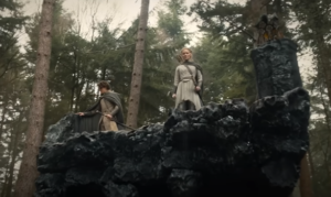 Shot from below looking up at Robert Aramayo as Elrond and Morfydd Clark as Galadriel, standing near the broken edge of an elevated stone walkway through a pine forest. Elrond has short tousled brown hair and wears a gray cloak over a pale yellowish-gray tunic. Galadriel has long blonde hair in a braid, and wears a gray cloak over a silver tunic with a quiver of arrows strapped to her back.