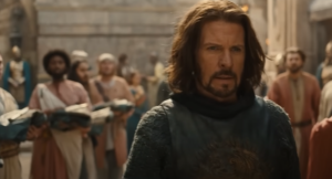 Lloyd Owen as Elendil, walking down a street between rows of people holding bundles of fabric in their arms. He has shoulder-length shaggy brown hair and a beard, and wears a faded blue tunic with a gold motif over a fish scale-patterned long-sleeved blue shirt.