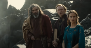 (left to right) Trystan Gravelle as Pharazon, Will Keen as an unnamed Numenorean scribe, and Ema Horvath as Earien, standing on a rocky shore looking off to the right with surprised expressions. Pharazon has long curly gray hair and a gray beard streaked with black and white. He wears a gold cape over the left shoulder of his red-and-gold robe. Will Keen's character has short brown hair, and wears a dark purple-brown mantle fastened with a gold chain across the front. He holds a large book under one arm. Earien has long brown hair in loose ringlets, and wears a dark blue mantle fastened by a gold chain over a dark blue dress.