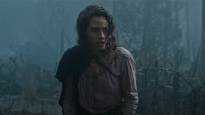 Nia Towle as Estrid, standing in a forest. She has long curly dark brown hair falling around her face, and wears a dark purple cape over the right shoulder of her faded pink blouse. She is standing in a forest.