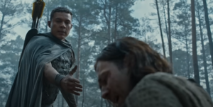 Ismael Cruz Cordova as Arondir, standing over Maxim Baldry as Isildur, extending a hand to the man. Arondir has close-cropped dark hair and wears a gray cloak over a gray wooden breastplate sculpted into the glowering face of a man with a leafy beard and hair. He has a quiver of arrows strapped to his back. Isildur has shoulder-length shaggy brown hair and wears a gray cloak. They are in a forest.