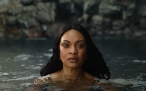 Cynthia Addai-Robinson as Miriel, standing in water up to her shoulders. She has long dark hair, and wears a white gown with gold brooches pinned on both shoulders.