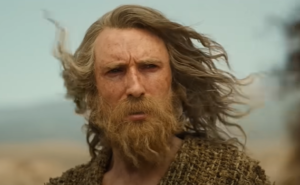 Daniel Weyman as the Stranger. He has long gray hair, whipping in the wind, and a short beard that appears more brown than gray. He is wearing a mossy gray-green robe.