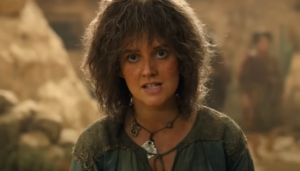 Markella Kavenagh as Elanor Brandyfoot in The Rings Of Power. She has short, curly brown hair, and wears a dark green blouse. A disc of silver hangs on a cord around her neck.