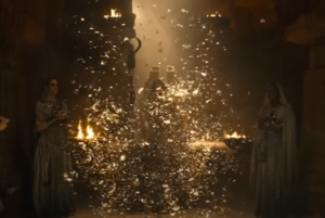 An underground temple with sandstone pillars. Women wearing white veils and floor-length gowns stand in the shadows, watching as a figure emerges from a swarm of brown butterflies and lifts a staff.