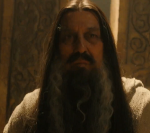 Ciaran Hinds as a wizard. He has long dark brown hair, very straight and parted in the middle, and a long beard streaked with black and white. He is wearing white robes.