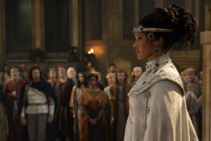 Cynthia Addai-Robinson as Miriel in The Rings Of Power, facing left, walking past a row of onlookers in a great hall. She has curly dark hair, bunched up and hanging in loose ringlets. She wears a silver diadem encrusted with large black gemstones, and a black-and-white mosaic collar over a long white gown.