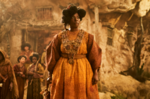 Tanya Moodie as Gundabel in The Rings Of Power, standing in the center of a village built into the steep walls of a sandy ravine. She has dark frizzy hair, and wears a saffron-yellow dress with wide reddish-brown sleeves and a long necklace made of knitted yarn, from which hangs a large round medallion.